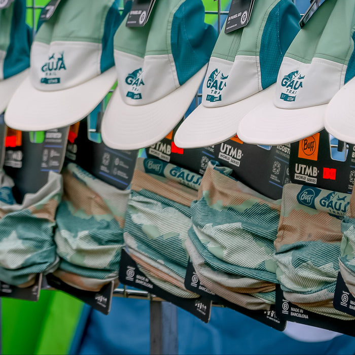 Multiple matching baseball caps hanging on a metal display rack, featuring a light blue and white landscape-style print on the front panels, tan brims, and teal side panels. The caps show the text 'AGA' and 'Menden' on the front, with small black brand tags attached.