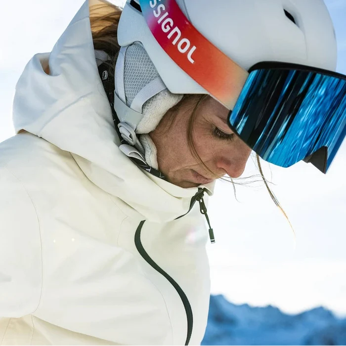 Rossignol ski goggles shown on a model in a snowy outdoor setting on the left, and a clean product rendering of the goggles on the right. The goggles feature a white helmet, blue mirrored lenses, and a strap with a red-to-purple gradient and the Rossignol logo.