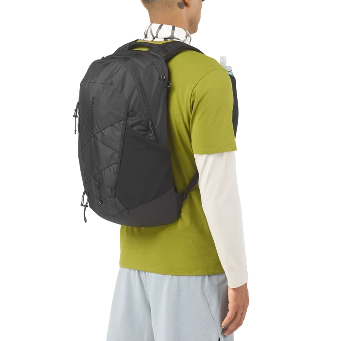 A man shown from the back wearing a black HOKS backpack, styled with a green and white long-sleeve shirt and light blue pants against a white background.